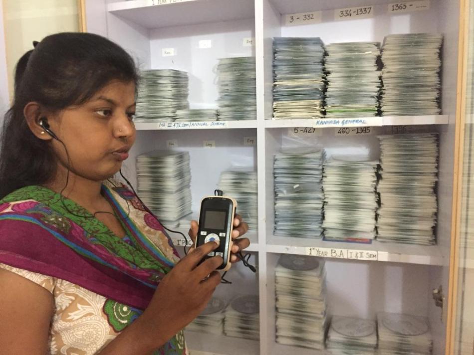 Mitra Jyothi beneficiary listening to Audio Books on a DAISY Player. Seen behind her is the Library's Master CD collection