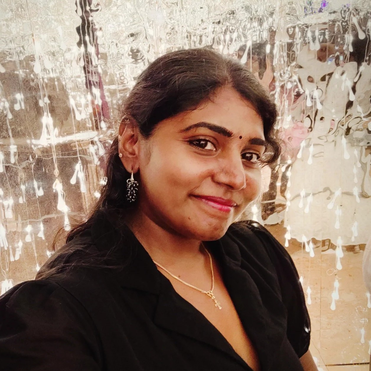 A woman smiling for a selfie in front of a reflective backdrop with bright, hanging light decorations.