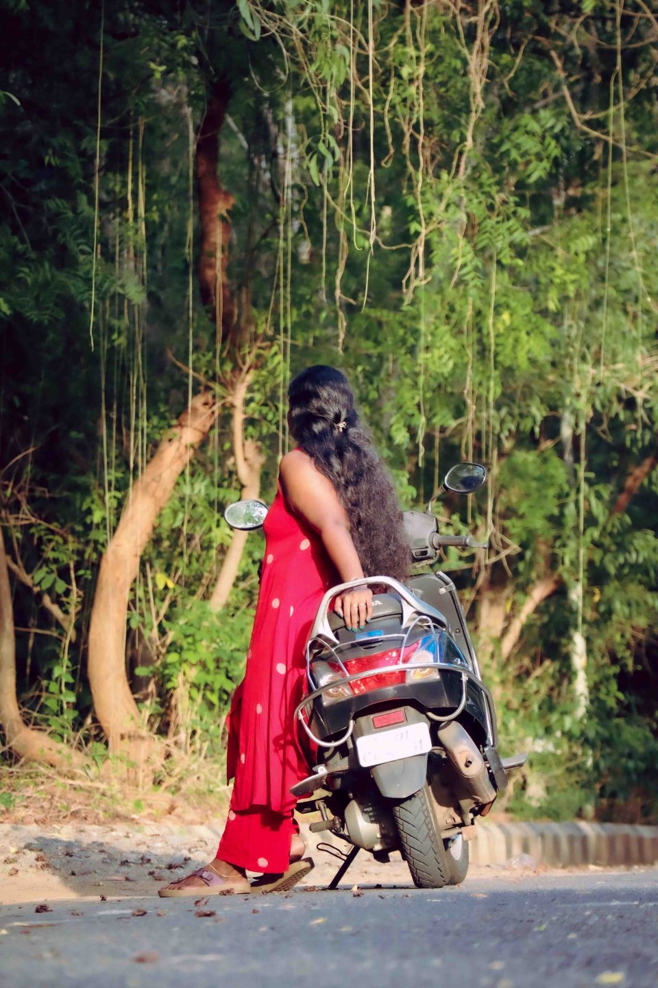 A person in a red dress with polka dots standing beside a motorcycle on a tree-lined road.